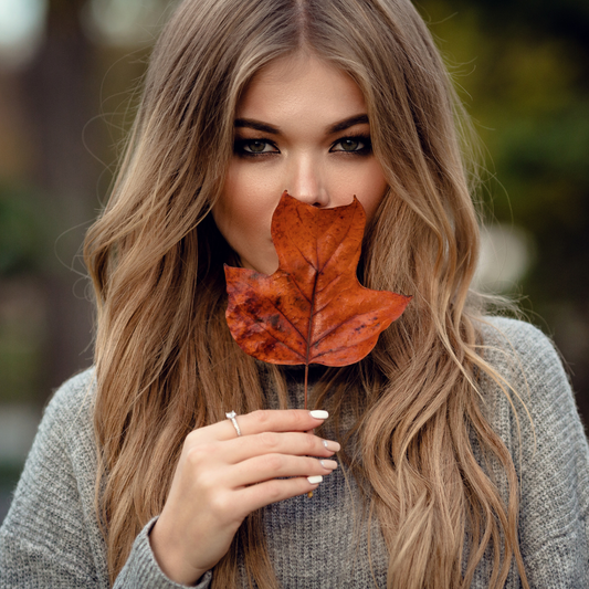 Beautiful woman with long blonde hair holding a fall leaf in front of her face, showcasing soft waves and autumn-inspired beauty — Bombshell salon and suites troy ny seasonal campaign.