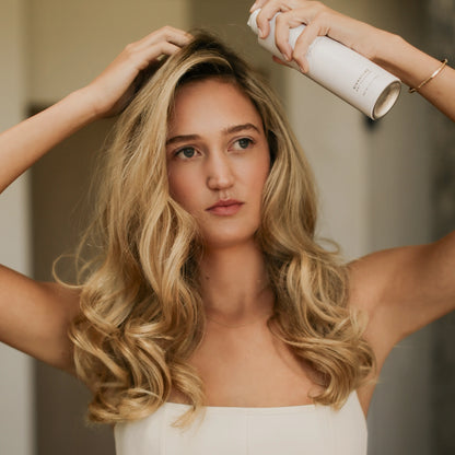 A woman with long, wavy blonde hair applies hairspray from a white aerosol can to her hair, wearing a white strapless top in a softly lit indoor setting.
