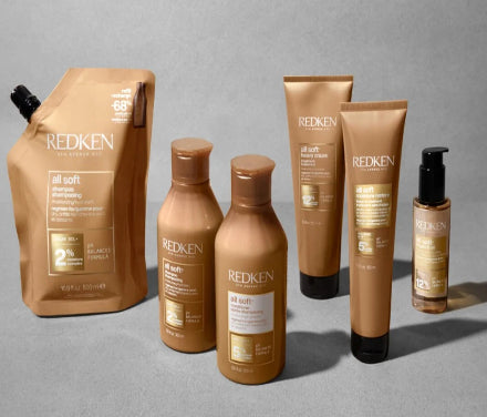 A collection of REDKEN All Soft haircare products in a coordinated metallic gold and bronze colorway, including a shampoo pouch, two shampoo bottles, two conditioner tubes, and a small treatment bottle, arranged on a neutral gray background.
