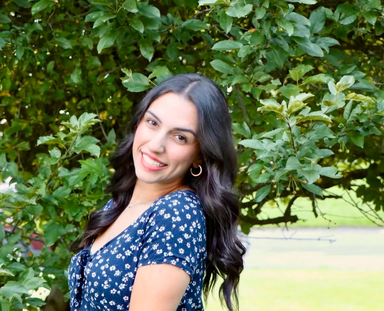 Woman in a blue floral dress standing in a park with trees and grass in the background